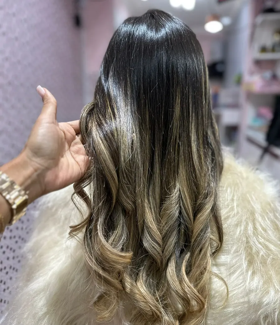 Cabelo castanho com mechas iluminadas e ondas largas, ajeitado pela mão da cabeleireira